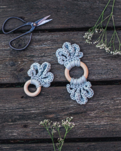 Two different sized teal coloured LoLo Loop Baby teethers are placed on a dark wooden backdrop with two flowers and a scissor framing them