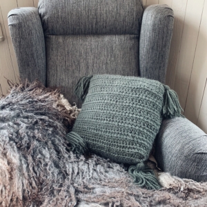 A moss green Stay Awhile Boho Pillow is placed in an armchair