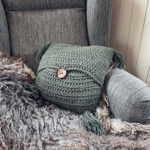 A moss green Stay Awhile Boho Pillow placed in an armchair